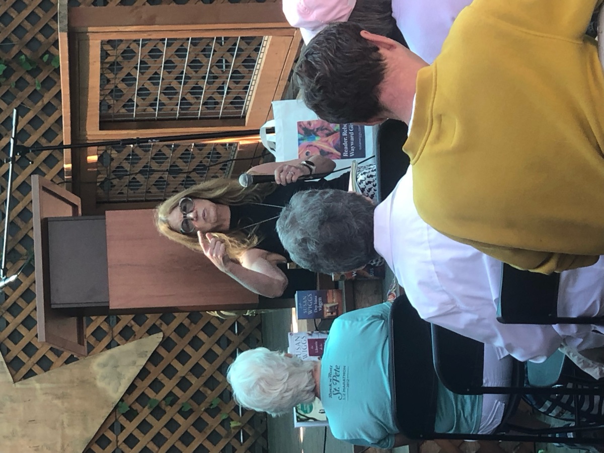 An author speaking into a microphone to a seated audience, with books on the stage behind her.