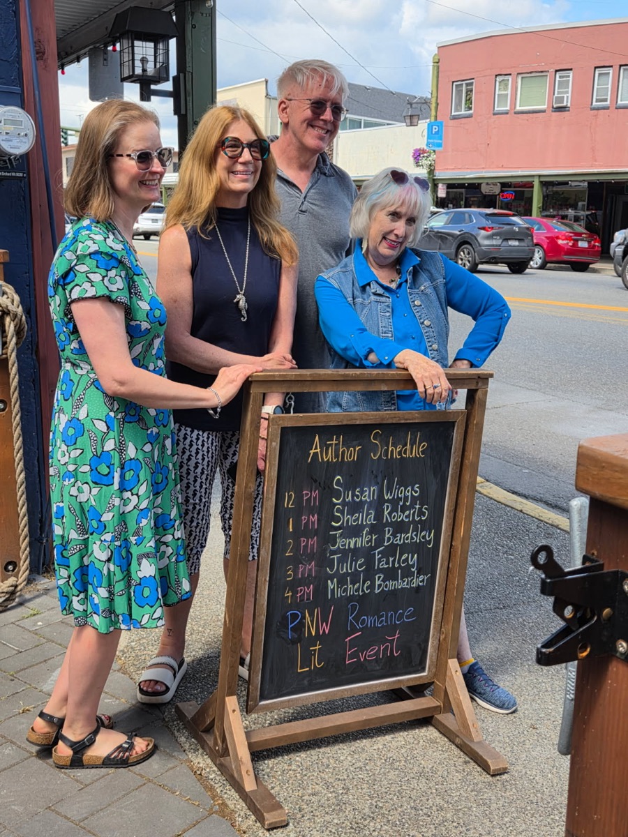 Four people smiling beside a chalkboard sign that lists an author reading schedule.